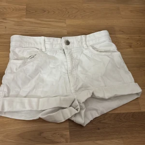 Vita jeansshorts - Säljer ett par vita shorts i denim med upprullad kant och klassisk femficksdesign. De har bälteshällor och stängs med knapp och dragkedja. Perfekta för varma dagar och enkla att matcha med olika toppar.