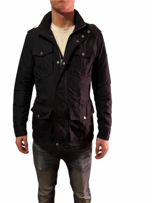 Svart vindjacka med dragkedja - Hålen vid fickorna syns inte vid användning och går att fixa hos en skräddare väldigt enkelt och billigt! Säljer en svart vindjacka med flera fickor framtill och snygga detaljer som knappar och dragkedja. Jackan har hög krage, axelklaffar och justerbar midja med snörning. Perfekt för dig som vill ha en stilren och funktionell jacka till vardags.