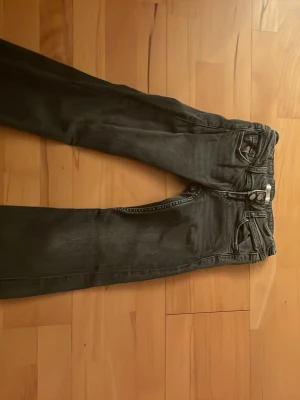 Svarta jeans med utsvängda låg midja det - Snygga svarta jeans med utsvängda ben och klassisk femficksdesign. Jeansen har svarta färg knappar på bakfickorna och är tillverkade i ett mjukt denimtyg. Perfekta för en stilren och avslappnad look.