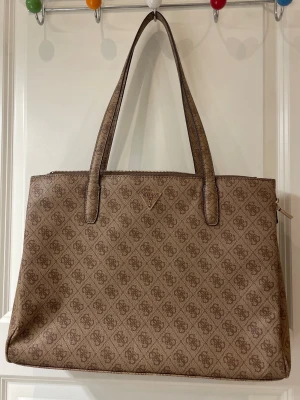 Beige handväska från Guess - Stilren beige handväska från Guess med klassiskt monogrammönster över hela väskan. Väskan har dubbla handtag, dragkedja upptill och en praktisk ytterficka med dragkedja på baksidan. Perfekt för dig som gillar snygga och rymliga väskor.