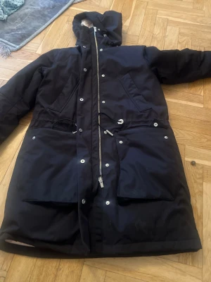 Svart parkas med huva och snörning M - Snygg svart parkas med huva, dragkedja och tryckknappar framtill. Jackan har stora fickor, snörning i midjan och en längre passform som skyddar mot kyla. Perfekt för kalla dagar och enkel att styla till olika outfits.