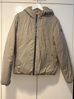 Beige dunjacka från Save The Duck - Snygg beige dunjacka från Save The Duck med huva och dragkedja framtill. Jackan har minimalistisk design, två sidofickor med dragkedja och logga på ärmen. Perfekt för kyliga dagar och enkel att matcha med olika outfits.