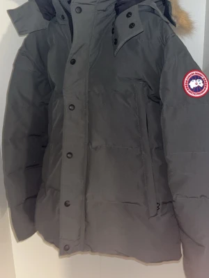 Grå dunjacka från Canada Goose - Snygg grå dunjacka från Canada Goose med pälskantad huva och klassisk logga på ärmen. Jackan har quiltad design, dragkedja och knappar framtill samt långa ärmar. Perfekt för kalla dagar och riktigt varm. Köpt begagnad för 8000kr. Har valt att sälja den för 4000kr för att köpa en lite billigare jacka. Kan diskutera pris i dm.