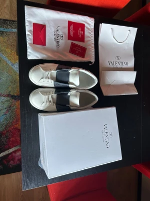 Valentino Garavani Open sneakers - Snygga vita Valentino Garavani Open sneakers med bred svart rem över mitten. Klassisk rund tå, vita snören och diskret logga på plösen. Skorna är i skinn och kommer med originalkartong, dustbag och tillbehör. De är använda fåtal gånger och är väl omhändertagna ‼️priset kan diskuteras ‼️