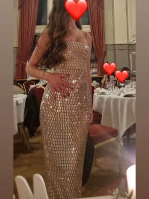 Glittrig långklänning i guld/champange - Säljer en glittrig långklänning i guld med figurnära passform och axelbandslös design. Band följer med som man kan sätta dit om man vill.  Klänningen har ett snyggt kristallmönster som reflekterar ljuset och ger en lyxig känsla. Perfekt för dig som vill sticka ut på bal eller fest.  Från oh polly använd endast en kväll. 