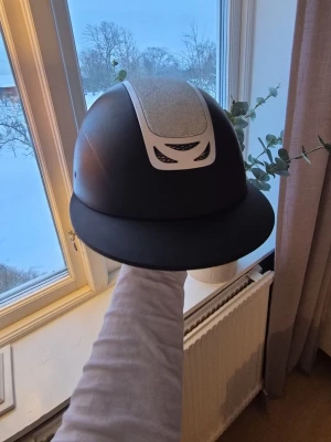 Svart ridhjälm Horze HZ Equestrian - Säljer en svart ridhjälm från Horze, modell HZ Equestrian Div. Hjälmen har ventilerande meshdetaljer och en stilren design med diskreta sömmar. Har lite repor på sidorna. Perfekt för dig som rider och vill ha både säkerhet och snygg look. Glittret är blå/silvrigt.💙