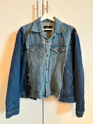 Patchwork jeansjacka i två blå toner - Unik jeansjacka från Weekday med patchwork-design i två olika blå nyanser. Jackan har råa kanter, klassisk krage, bröstfickor med knappar och knappstängning framtill. Oversized passform för en avslappnad look. Perfekt för dig som vill sticka ut. Storlek: M/L