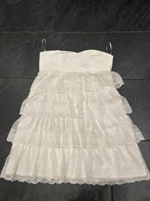 Bandeau studentklänning - Jätte söt vit kortklänning med tunna axelband och flera lager volanger i spets. Klänningen har en dragkedja i ryggen och är perfekt för sommarens alla tillfällen. Romantisk look med fina spetsdetaljer och feminin passform. Funkar perfekt som studentklänning! 