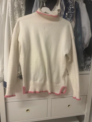 Vit stickad polotröja med rosa detaljer - Vit stickad polotröja med ribbade muddar och rosa detaljer vid hals, ärmslut och nederkant. Tröjan har en avslappnad passform och hög krage, perfekt för lager-på-lager-stil.