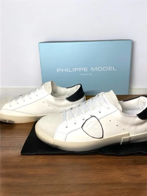 Philippe Model vita sneakers - Stilrena vita sneakers från Philippe Model med svarta detaljer vid hälen och logotyp på sidan. Skorna har snörning, rund tå och är tillverkade i skinn med inslag av mocka framtill. Perfekta för en clean och modern look.