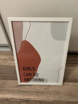 Tavla med text 'Girls Can Do Anything' - Tavla med vit ram och abstrakt motiv i beige, rött och grått. Texten 'Girls Can Do Anything' tryckt på framsidan. Själva bandet som ska sitta på baksidan mellan ramen och ”tavelfoten” är borta. Inget som påverkar hur tavlan står upp. En klisterlapp är lite kvar på ramens baksida. 