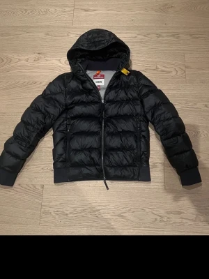 Parajumpers Jacke Sheen - Snygg svart dunjacka från Parajumpers, modell Sheen. Jackan har huva, quiltad design och dragkedja framtill. Detaljer som gul rem vid halsen och ribbade muddar vid ärmslut. Perfekt för kalla dagar och riktigt stilren look.
