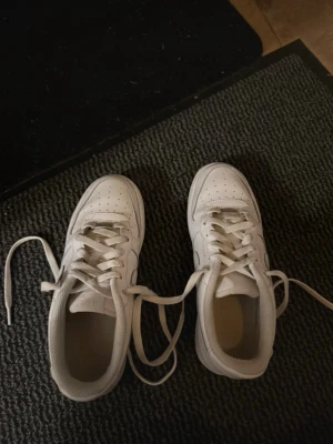 Vita Nike Air Force 1 sneakers - Klassiska vita Nike Air Force 1 sneakers med låg profil och rund tå. Skorna har snörning och är tillverkade i skinn med perforerade detaljer på tån för extra ventilation. Perfekta för dig som gillar en clean och tidlös stil.