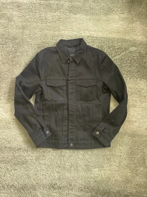 Svart denimjacka från J.Lindeberg - Rikitgt snygg och stilren svart denimjacka med raka linjer och två bröstfickor med lock. Jackan har klassisk krage, knappar framtill och vid ärmslut. Perfekt passform. Vid funderingar, fråga på! 