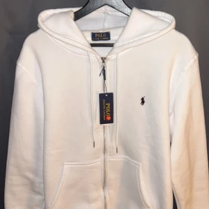 Vit hoodie från Polo Ralph Lauren - Vit hoodie från Polo Ralph Lauren med dragkedja framtill och klassisk huva. Tröjan har två fickor och den ikoniska broderade loggan på bröstet. Perfekt för en clean och stilren look.