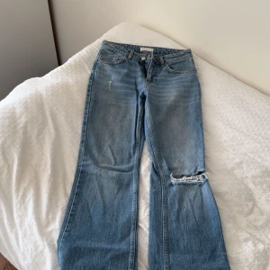 Blå bootcut jeans från Perfect Jeans - Snygga blå bootcut jeans från Perfect Jeans i storlek 34. Jeansen har en låg midja, slitningar och hål på ena knät för en trendig look. Tillverkade i klassiskt denimtyg med fem fickor och dragkedja. Perfekta för dig som gillar en avslappnad och cool stil.