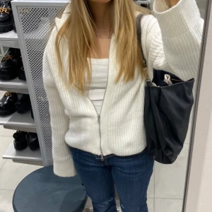 Vit ribbad kofta med dragkedja - Mysig vit kofta i ribbstickat material med dragkedja framtill. Koftan har lång ärm och en avslappnad passform, perfekt att slänga över ett linne eller t-shirt. Enkel och stilren design som passar till jeans eller kjol.