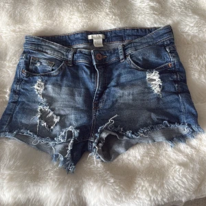 Blå jeansshorts med slitningar från H&M - Snygga blå jeansshorts från H&M i storlek 38. De har råa, fransiga kanter och flera slitningar både fram och bak för en cool, avslappnad vibe. Klassisk femficksmodell med knapp och dragkedja. Perfekta för sommaren och festivalhäng. Väldigt lågmidjade 