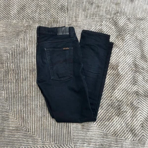 Svarta jeans från Nudie  - Svarta jeans från Nudie Jeans i klassisk femficksmodell. Jeansen har en slim passform med raka ben och är tillverkade i ett mjukt och stretchigt bomullsmaterial. Snygga detaljer med svarta sömmar och diskret logga på bakfickan. Perfekta för dig som gillar stilrena och bekväma jeans.