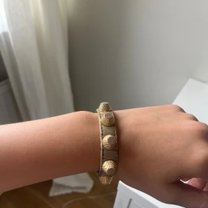 Balenciaga armband med guldnitar - Snyggt beige armband i skinn med stora, texturerade guldnitar och spänne i guld. Armbandet har en cool och edgy vibe, perfekt för att lyfta vilken outfit som helst. Justerbar längd med flera hål för bästa passform.