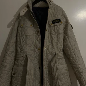 Beige quiltad jacka från Barbour - Quiltad beige jacka från Barbour med guldiga knappar, dragkedja och bälte i midjan. Jackan har hög krage, flera fickor och Barbour International-märke på bröstet. Super snygg!!