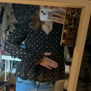 Svart blommig blus med volang - Säljer en svart blus med små blommiga mönster i beige. Blusen har volangdetaljer vid ärmslut och nederkant, samt långärmade transparenta ärmar. V-ringad hals med knytband och en luftig, loose passform. Perfekt för dig som gillar en söt och trendig stil.