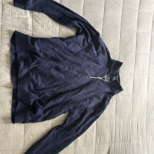 Mörkblå Zip Tröja - Mörkblå sweatshirt med half zip och hög krage. Tröjan har långa ärmar och ribbade muddar vid ärmslut och nederkant. Perfekt för lager-på-lager och chill dagar. Materialet känns mjukt och bekvämt, troligtvis bomull eller bomullsblandning..
