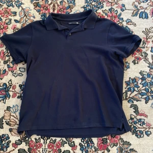 Mörkblå pikétröja från 157 - Snygg mörkblå pikétröja från 157 i storlek L. Klassisk krage och korta ärmar, tillverkad i mjuk bomull. Perfekt för dig som gillar enkel och stilren design. Tröjan har två knappar vid halsen och en rak passform.