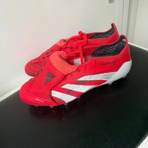 Adidas Predator röda fotbollsskor - Säljer ett par röda Adidas Predator fotbollsskor med vita och svarta detaljer. Skorna har snörning, klassisk design med täckt plös och dobbar för gräsplan. Snygga vita ränder på sidorna och Predator-tryck undertill. Perfekta för dig som vill sticka ut på planen.