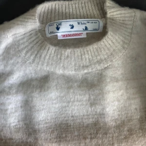 Off-White stickad tröja i beige och grå - Säljer en stickad tröja från Off-White med ikoniska pilar på ryggen. Tröjan har en snygg gradient från beige till mörkgrå och är långärmad med ribbad hals. Materialet är mjukt och fluffigt, perfekt för dig som gillar streetwear med lyxig känsla. Storlek S men passar M också.