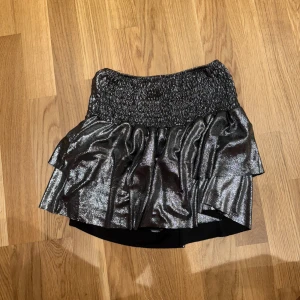 Glittrig svart volangkjol Meet x Tiger - Säljer en glittrig svart kortkjol från Meet x Tiger med bred smock i midjan och dubbla volanger. Kjolen har en metallic finish som verkligen sticker ut och ger en cool vibe. Perfekt för dig som vill ha något extra till outfiten!