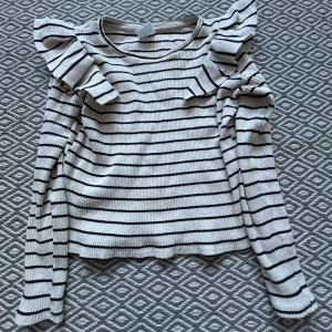 Randig vit tröja med volang från Zara - Snygg vit långärmad tröja från Zara med svarta ränder och coola volangdetaljer på axlarna. Ribbad struktur och rund hals. Perfekt för dig som gillar en trendig still. Passar till mycket och storleken är 140