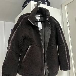 Mörkbrun teddyjacka från H&M XXS - Den virala jackan, slutsåld som vanligt! Helt ny.  Mysig svart teddyjacka från H&M i storlek XXS. Jackan har hög krage, dragkedja framtill och detaljer i skinnimitation längs ärmar och fickor. Perfekt för kalla dagar och ger en cool vibe till din outfit.