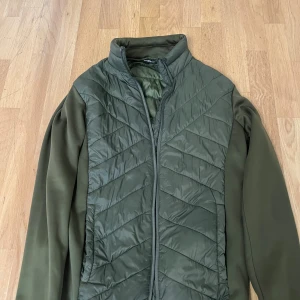 Olivgrön quiltad pufferjacka - Snygg olivgrön pufferjacka med quiltad framsida och rygg. Jackan har lång ärm, hög krage och dragkedja framtill. Materialet är en mix av polyester och fleece, vilket gör den både lätt och varm. Perfekt för kyliga dagar. Perfekt nu till hösten(: