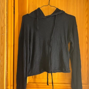 Svart ribbad hoodie från Gina Tricot - Svart ribbad hoodie från Gina Tricot i storlek XXS. Tröjan har långa ärmar, huva och snörning nertill. Mjuk och skön med en croppad passform, perfekt för en chill och avslappnad stil.