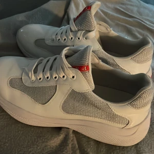 Prada American Cups  - Snygga vita sneakers från Prada med meshpaneler och detaljer i blankt syntetmaterial. Skorna har klassisk snörning och tydlig röd Prada-logga på plösen och innersulan. Fräscha och sportiga med chunky sula och modern design. 