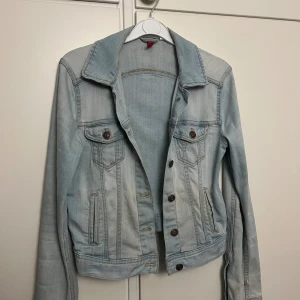Ljusblå jeansjacka med knappar - Snygg ljusblå jeansjacka med klassisk krage och bruna knappar framtill. Jackan har två bröstfickor med lock och knappar samt två sidofickor. Perfekt för lager-på-lager och enkel att styla till olika outfits.