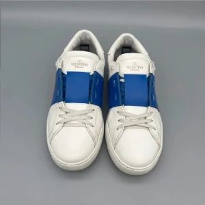 Valentino Garavani Open sneakers vit/blå - Snygga Valentino Garavani Open sneakers i vitt skinn med en bred blå rem över mitten. Klassisk rund tå, vita snören och coola nitar baktill på sulan. Perfekta för dig som vill sticka ut med en lyxig och modern look.