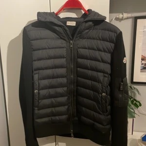 Svart Moncler Cardigan - Svart Cardigan från Moncler med stickade ärmar och quiltad front. Den är i bra skick nästan perfekt och det är inget fel på den. Jackan har huva, dragkedja framtill och fickor med tryckknappar. Klassisk Moncler-logga på ärmen och snygga detaljer som ger en sportig vibe. Perfekt för kyliga dagar. Fråga gärna om det är något och fler bilder går alltid att få