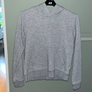 Grå hoodie från Gant med broderi - Snygg grå hoodie från Gant med broderad logga i silvertråd över bröstet. Tröjan har huva, magficka och långa ärmar. Perfekt för en chill och stilren look. Mjuk och skön bomullsblandning.