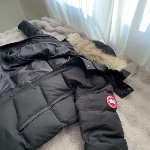 Svart dunjacka från Canada Goose - Säljer en svart dunjacka från Canada Goose med fluffig päls på huvan och klassisk logga på ärmen. Jackan har dragkedja, knappar och stora fickor framtill. Perfekt för kalla vinterdagar och riktigt varm med sin tjocka vaddering. Den är i jätte bra skick inga skador eller nåtnting som drabbar utseendet!