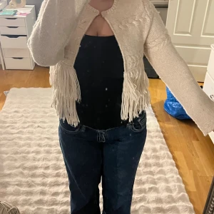 Beige stickad kofta med fransar Lindex - Superfin beige stickad kofta från Lindex i storlek S. Koftan är kortare i modellen, har flätstickade detaljer och coola fransar längs nederkanten. Perfekt för dig som vill ha en unik och trendig look med boho-vibe.