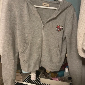 Grå hoodie från Zadig & Voltaire - Grå stickad hoodie från Zadig & Voltaire med dragkedja framtill och huva. Tröjan har ett broderat hjärta på bröstet och två fickor fram. Materialet är en mix av ull och polyamid, vilket gör den både mjuk och varm. Perfekt för kyliga dagar.