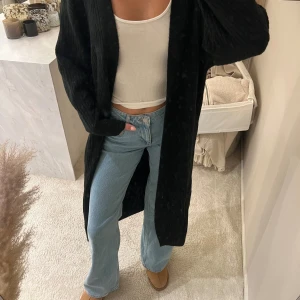 Svart lång stickad kofta från H&M - Säljer en svart, lång och ribbad stickad kofta från H&M. Koftan har en öppen front utan knappar eller dragkedja och är gjord i mjukt stickat material. Perfekt att slänga över en t-shirt eller topp för en chill look. Passformen är loose och koftan går ner till knäna.