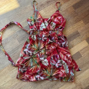 Klänning blommig  Guess höstfärger  - Säljer en färgstark kortklänning från Guess med tropiskt blommigt mönster i rött, grönt och beige. Klänningen har lite omlottmodell, smala axelband och ett knytband i midjan som framhäver siluetten. Perfekt för dagens väder med lite brittsommar. Är markerad Large. Men måste vara italiensk large, passar medium 
