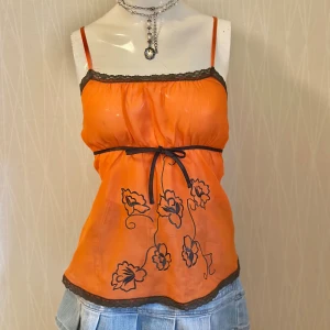 Orange blommig linnetopp med spets - Superfin orange linnetopp med svarta broderade blommor och tunna axelband. Toppen har svarta spetsdetaljer längs kanterna och ett smalt knytband i midjan. Materialet är lätt och transparent, perfekt för en trendig och färgstark look.