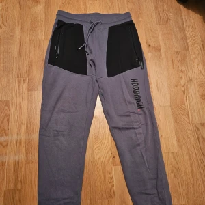 Grå Hoodrich mjukisbyxor med svarta detaljer - Snygga grå mjukisbyxor från Hoodrich med svarta paneler vid fickorna och svart logga längs benet. Byxorna har dragsko i midjan och dragkedjor på fickorna för extra stil och funktion. Perfekta för chill eller streetwear.