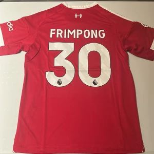 Liverpool Frimpong #30 matchtröja Adidas - Liverpool FC matchtröja i rött med vita detaljer, tryck Frimpong #30 på ryggen och Premier League-logga. Adidas Aeroready-material, kortärmad med klubbmärke och sponsortryck framtill. Perfekt för dig som älskar fotboll och vill sticka ut på läktaren. Replika, endast handtvätt gäller!