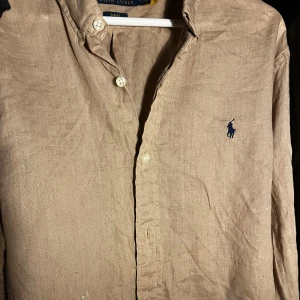 Beige linneskjorta från Polo Ralph Lauren - Säljer en beige långärmad skjorta från Polo Ralph Lauren i slim fit. Skjortan är tillverkad i linne och har klassisk krage, knäppning framtill och den ikoniska broderade loggan på bröstet. 