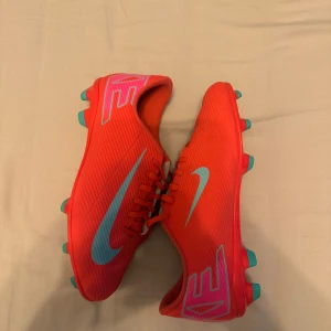 Nike fotbollsskor i rött och turkos - Säljer ett par Nike fotbollsskor i en riktigt snygg röd färg med turkosa detaljer och dobbar. Skorna har snörning och är designade för att ge bra grepp på planen. Perfekta för dig som vill sticka ut på matchen!